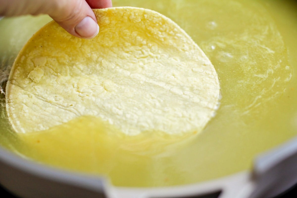 Corn tortilla being dipped in green enchilada sauce.